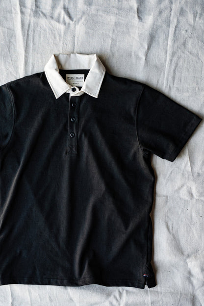 Short-sleeve basalt black rugby jersey with white collar and button placket, laid flat on linen.