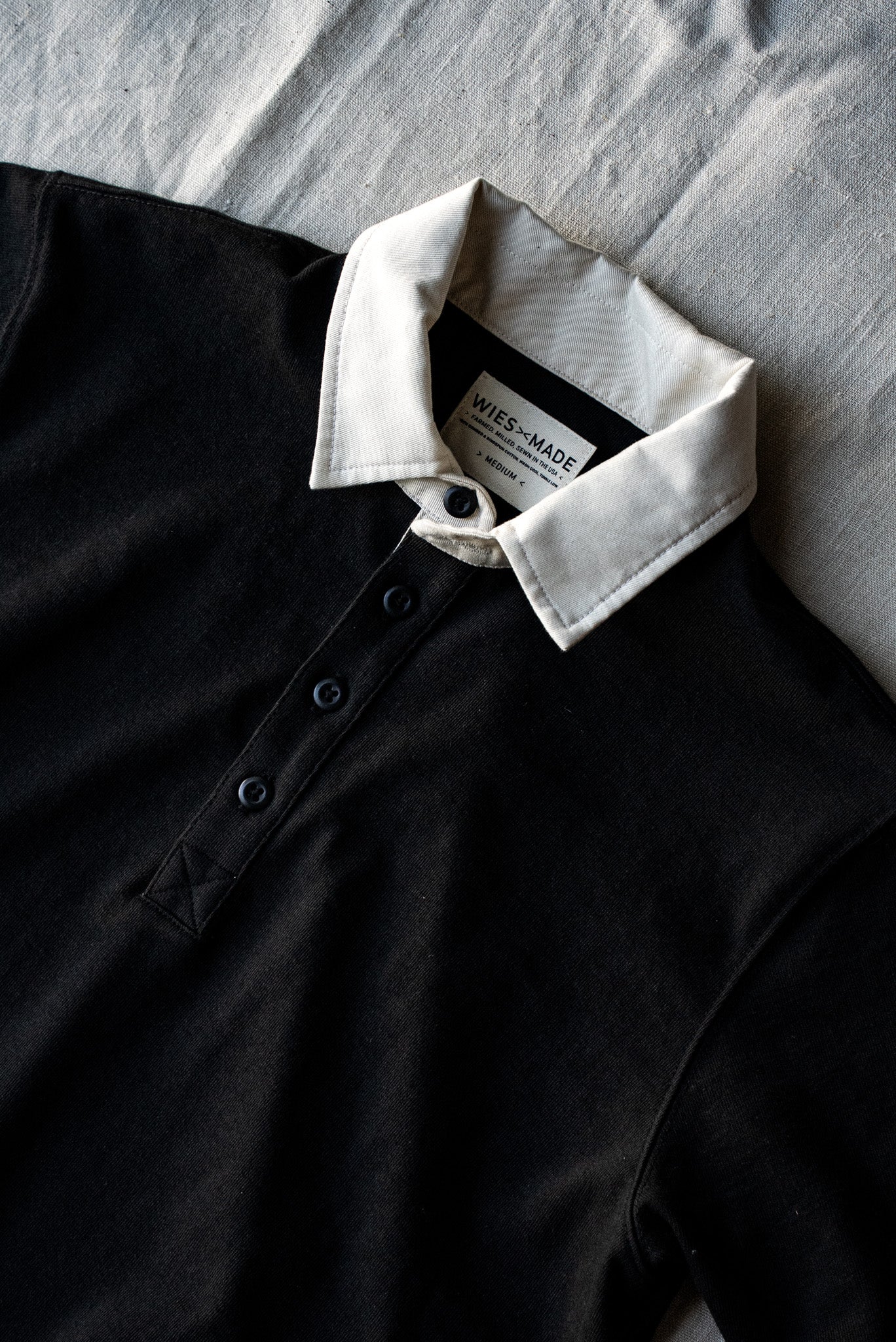 Close-up of black cotton rugby jersey with white twill collar, button placket, and WiesMade label.