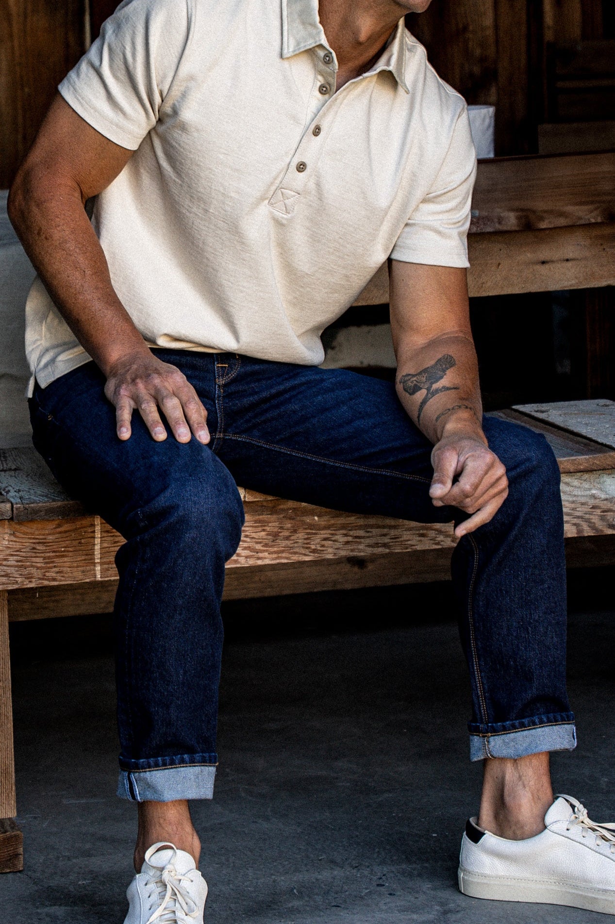 Man in natural rugby jersey polo and cuffed selvedge denim jeans seated on a wooden bench.