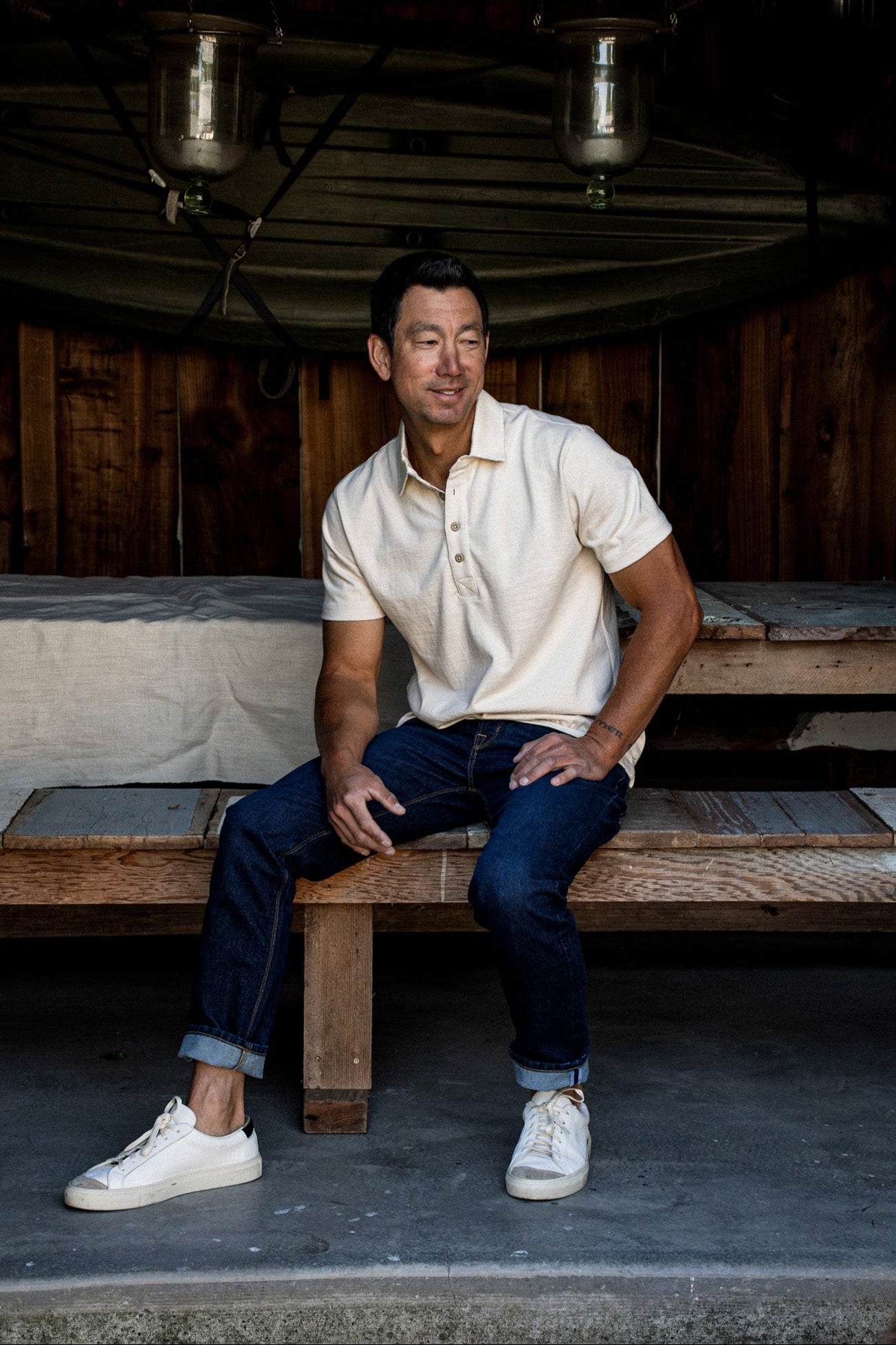 Man wearing a natural rugby jersey polo, seated on a wooden bench outdoors.