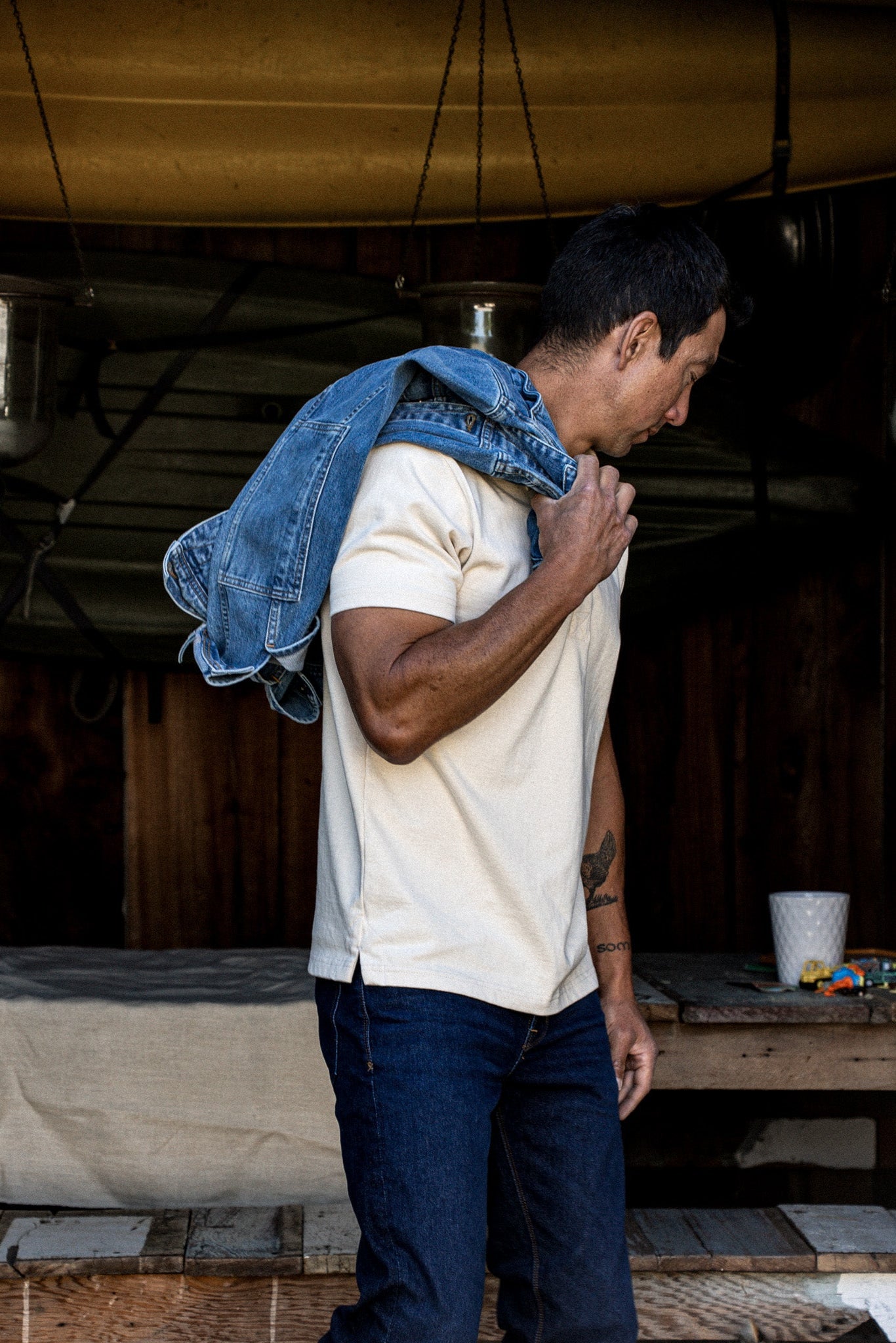 Side view of a man in a natural cream rugby shirt and dark denim jeans with a blue denim jacket over his shoulder.