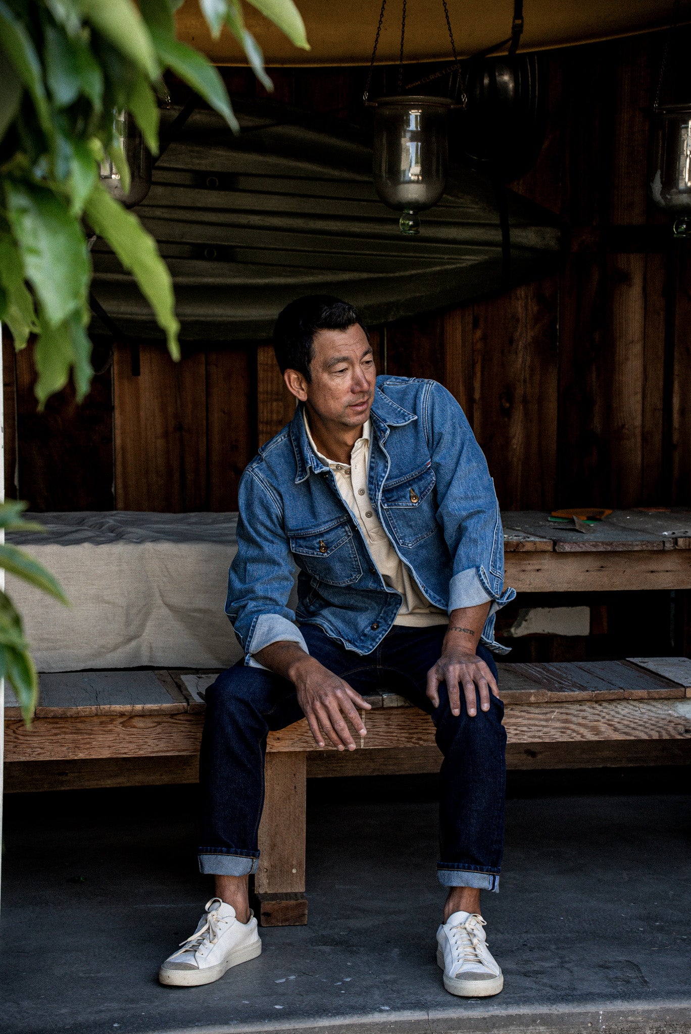 Man in cream cotton rugby jersey layered under a denim jacket, seated on a wooden bench.