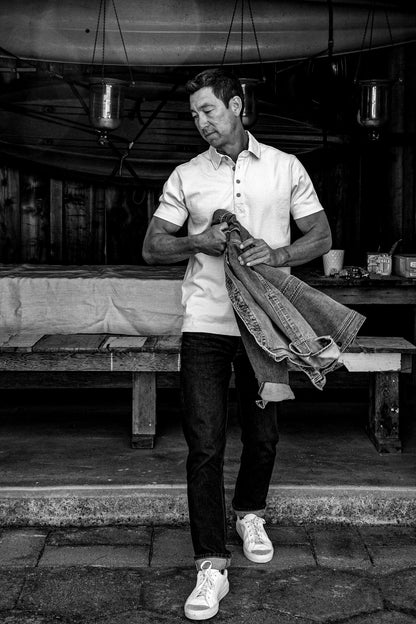 Man in a natural short-sleeve cotton rugby shirt holding folded raw denim jeans.