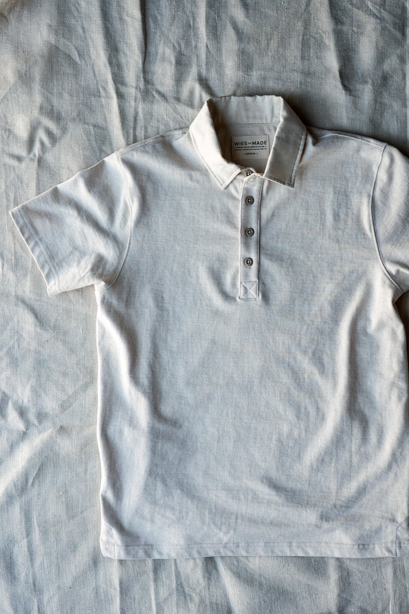Natural off-white short-sleeve cotton rugby jersey polo with three-button placket, laid flat on linen.