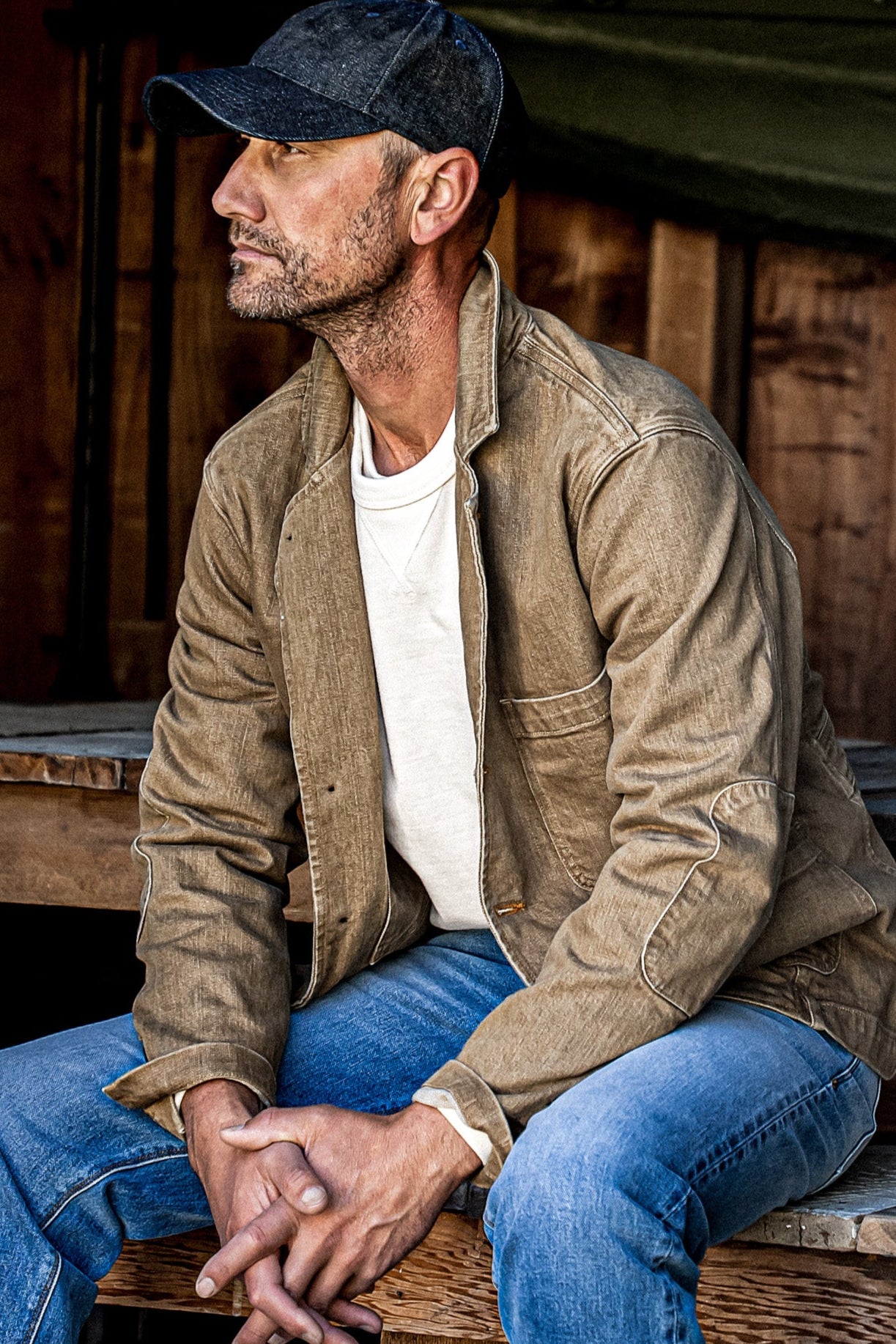 Man in wheat denim chore blazer over a white tee, seated against weathered wood.