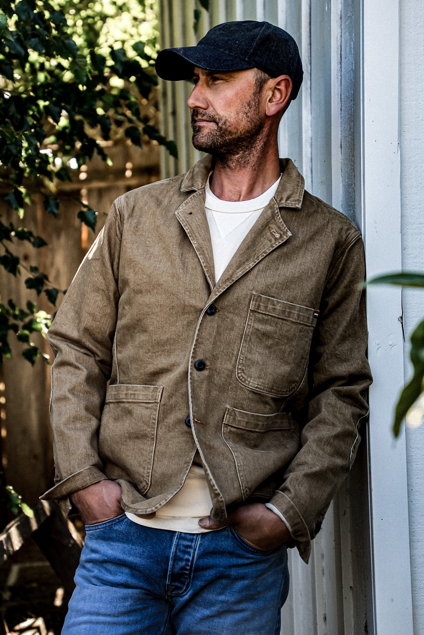 Man wearing wheat denim chore blazer with jeans, leaning against a white wall outdoors.