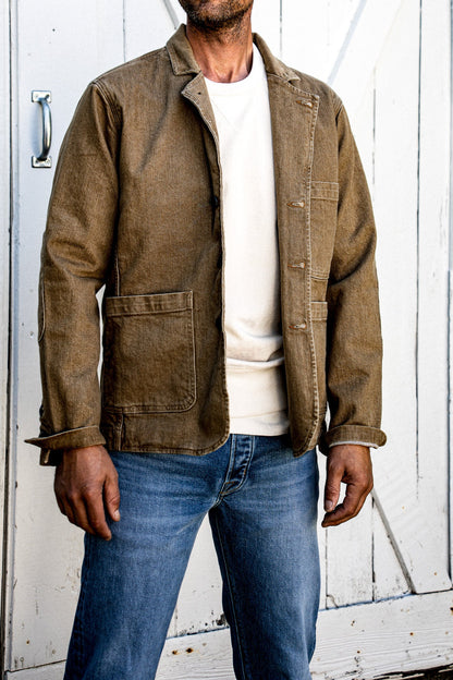 Man wearing wheat denim chore blazer over cream tee with jeans by white barn door.