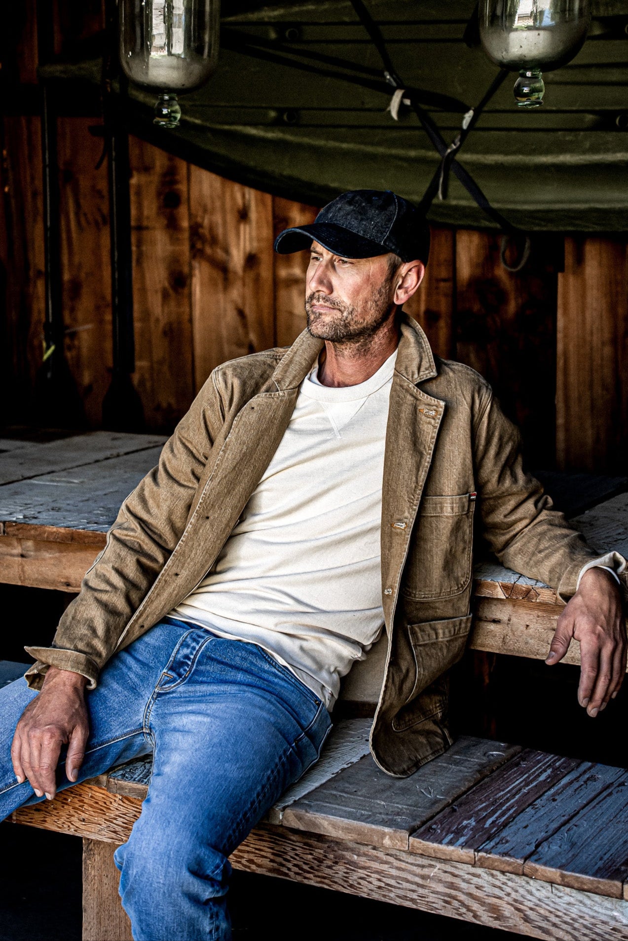 Man in wheat denim chore blazer over cream tee, jeans, and cap on rustic wood.