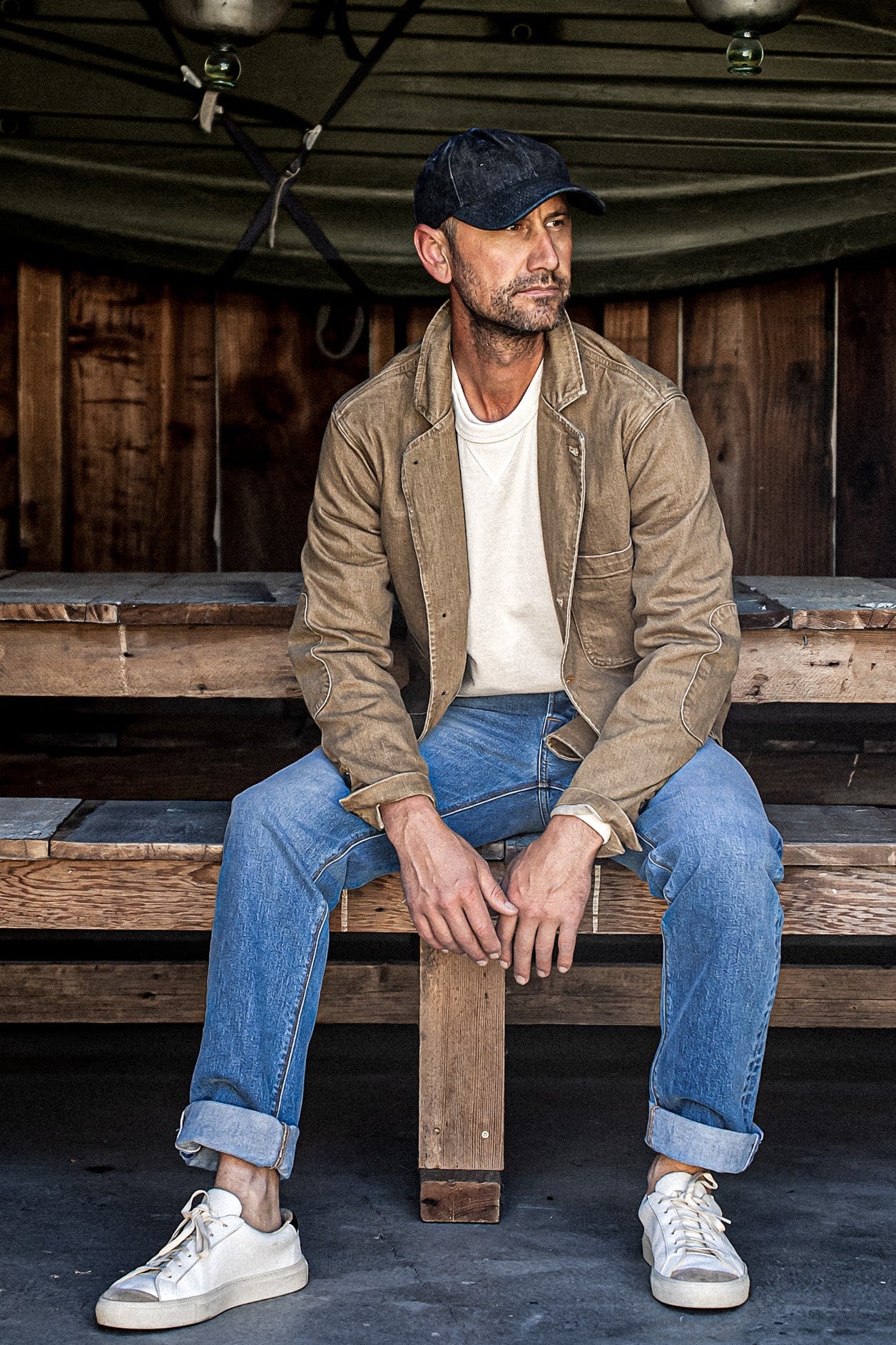 Man in wheat denim chore blazer over white tee and blue jeans on wooden bench.