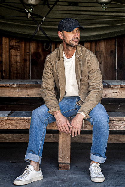 Man in wheat denim chore blazer over white tee and blue jeans on wooden bench.