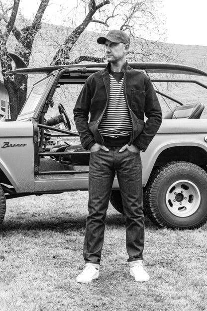 Man in black denim chore blazer, striped tee, and jeans standing beside a vintage Bronco.