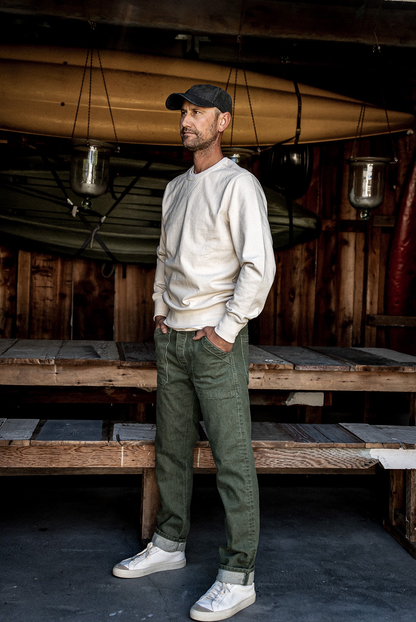 Man in olive Scout military denim pants and cream sweatshirt standing in a rustic boathouse.