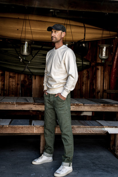 Man in olive Scout military denim pants and cream sweatshirt standing in a rustic boathouse.
