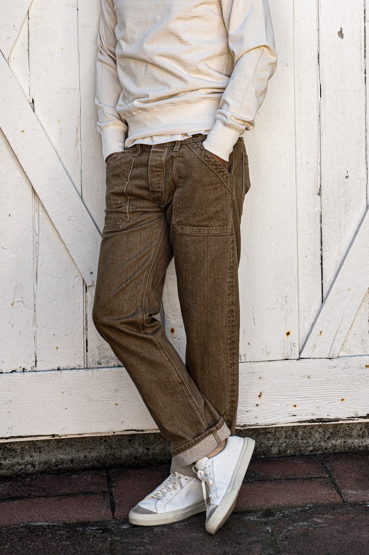 Wheat Scout military denim jeans worn on-model, leaning against a weathered white barn door.