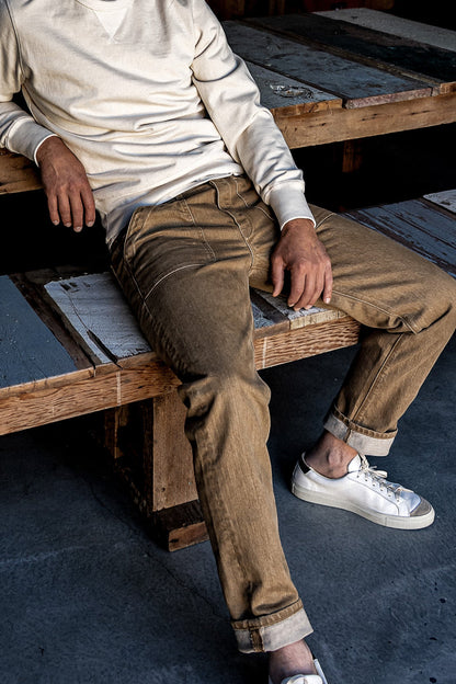 Person in wheat Scout military denim jeans with cuffed hems, seated on wooden pallets.