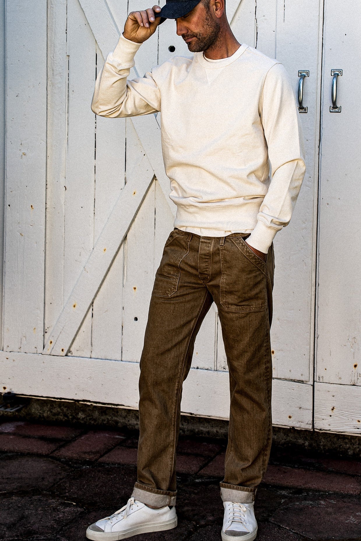 Man in wheat Scout military denim pants with cuffed hems, cream sweatshirt and cap.