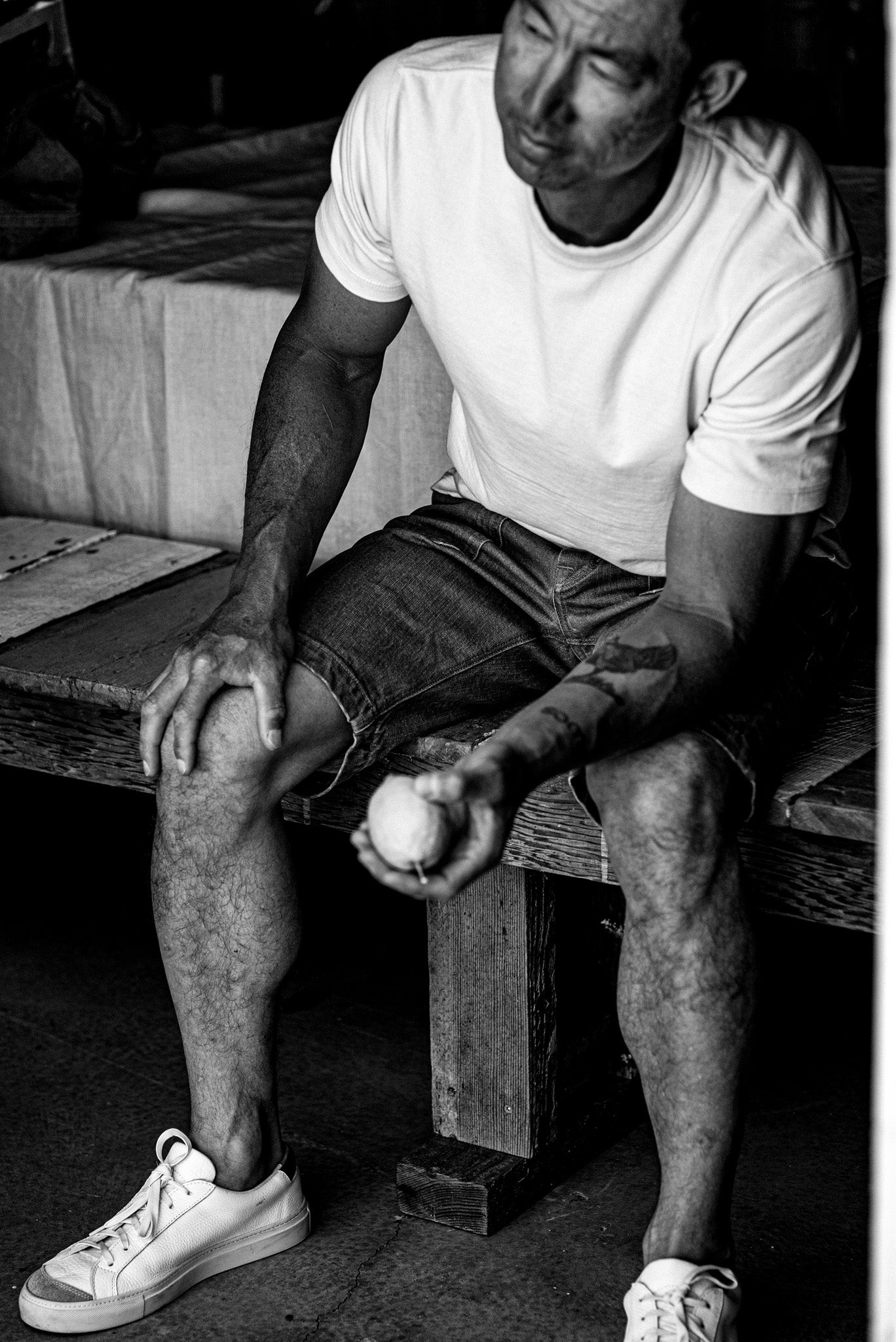 Man in white tee and broken twill denim military shorts seated on a wooden bench, holding a baseball.