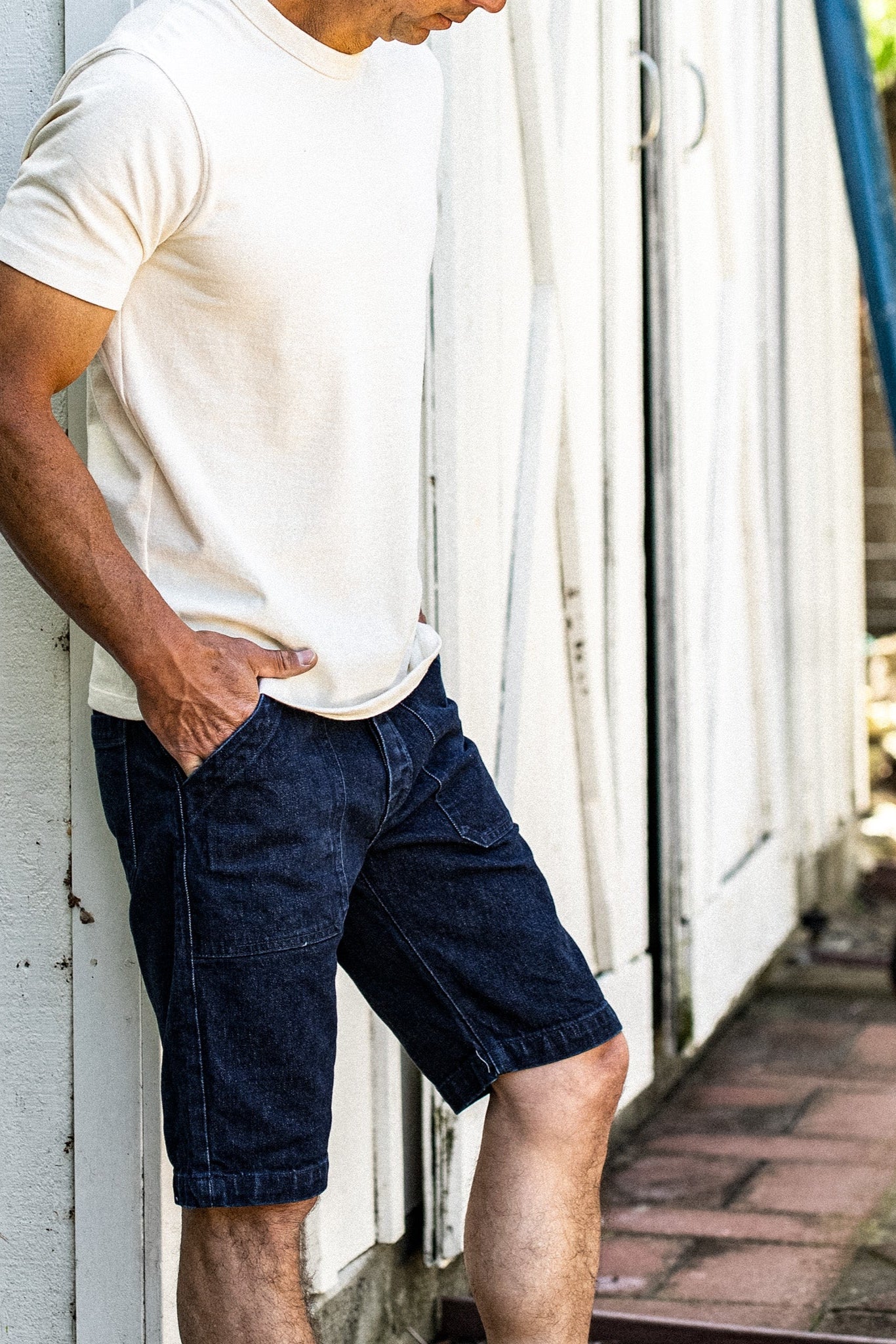 Man in cream tee and ocean wash denim military shorts leaning against a weathered white wall.