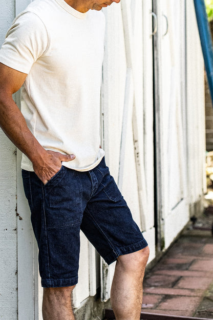 Man in cream tee and ocean wash denim military shorts leaning against a weathered white wall.