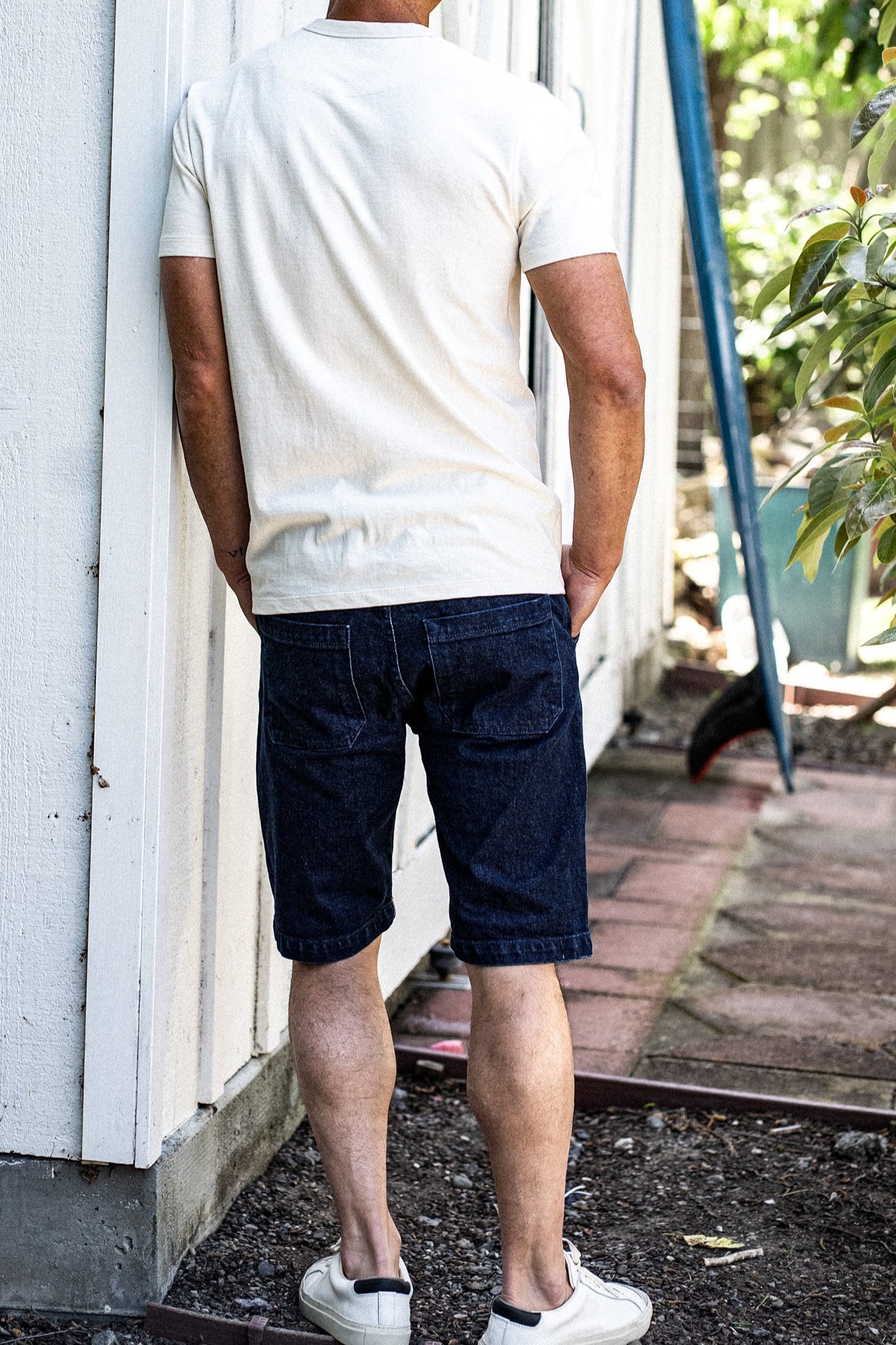 Back view of man in dark ocean wash denim military shorts and cream tee, leaning against a white wall.