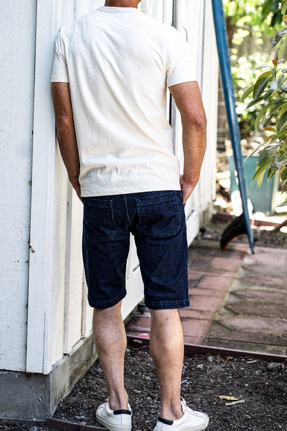 Back view of man in dark ocean wash denim military shorts and cream tee, leaning against a white wall.