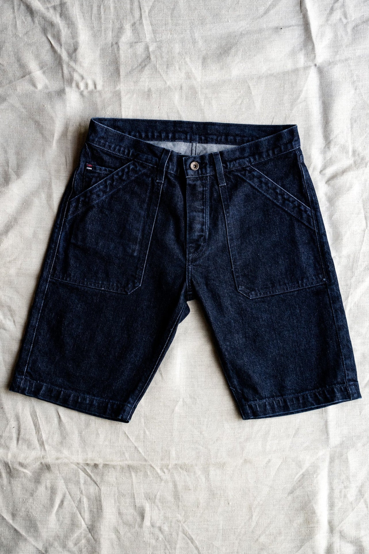 Dark ocean wash denim military shorts with patch pockets, laid flat on natural linen.