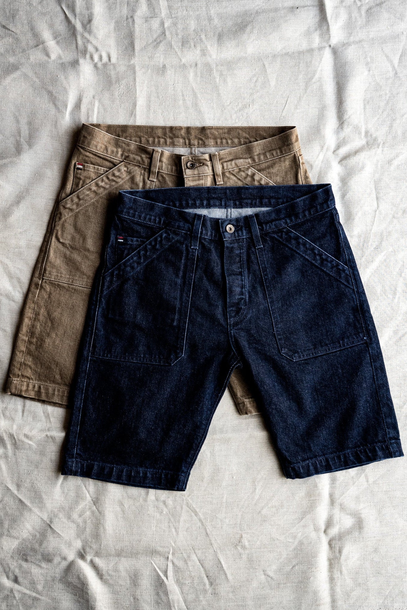 Dark indigo Scout denim military shorts layered over a khaki pair on linen, showing patch pockets.