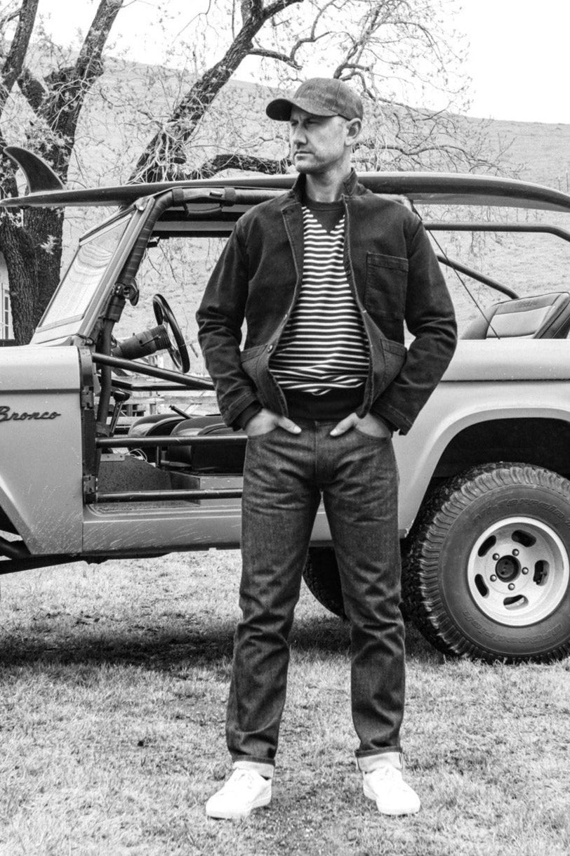 Man wearing a striped rugby jersey pullover under a jacket, standing by a vintage Bronco.