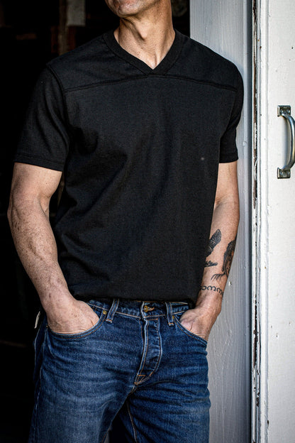 Man in dark Stinson V-neck cotton jersey and denim jeans leaning against a weathered door.