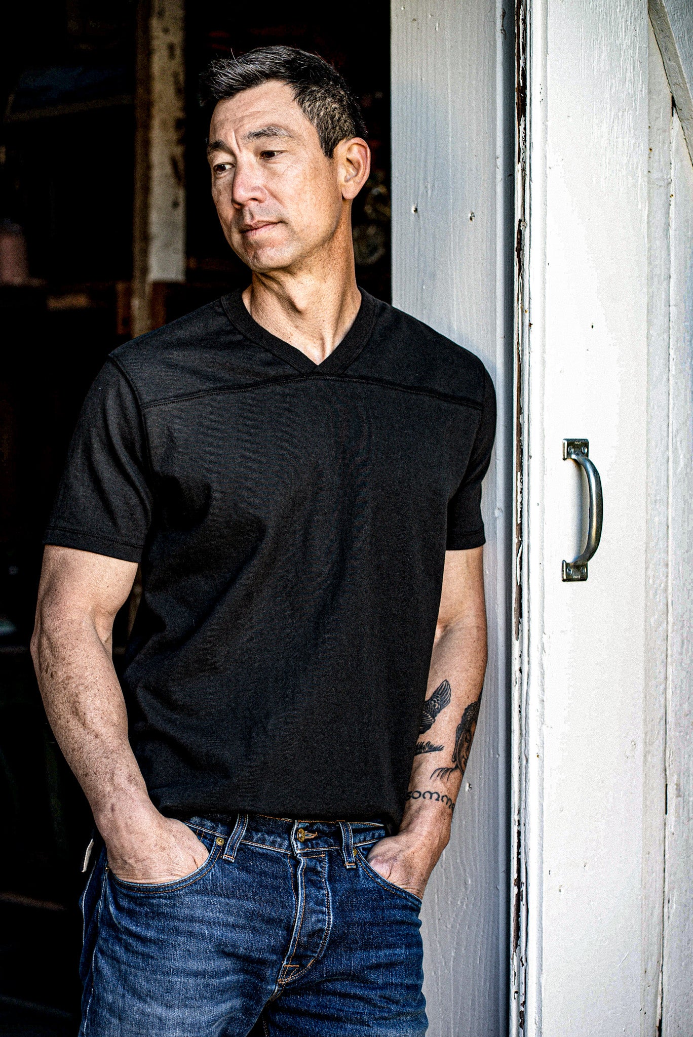 Man wearing basalt black Stinson V-neck jersey with denim jeans, leaning against a weathered door.