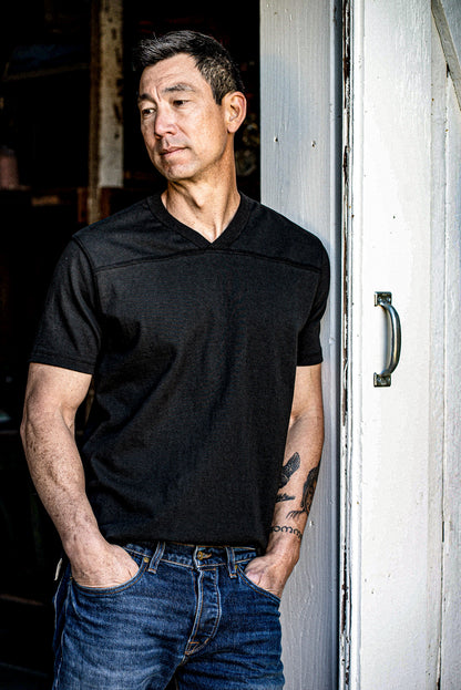 Man wearing basalt black Stinson V-neck jersey with denim jeans, leaning against a weathered door.