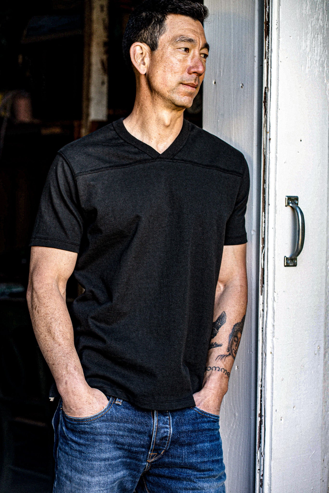 Man leaning against a weathered door in a black 12oz cotton V-neck jersey and jeans.