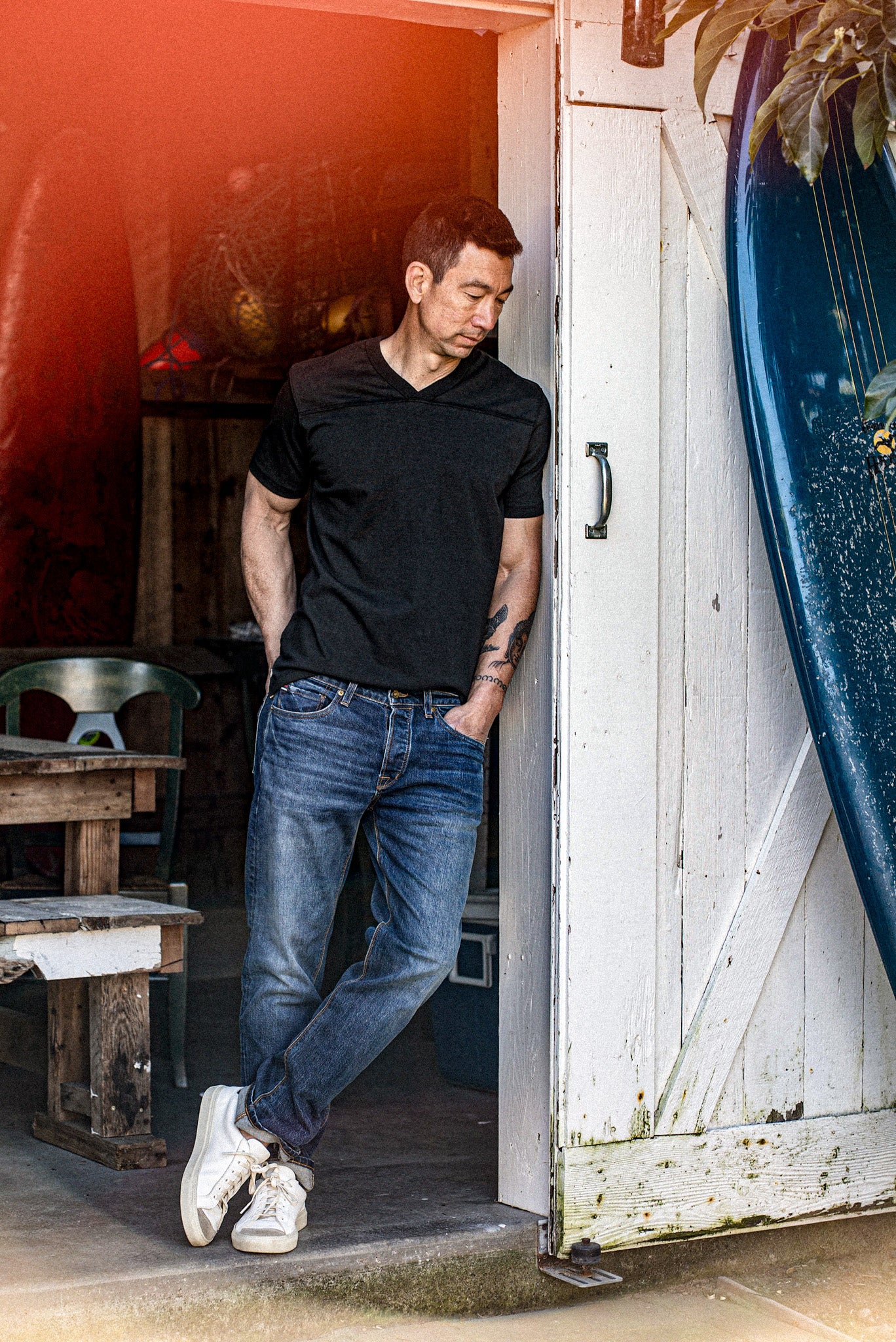 Man in black cotton V-neck jersey tee leaning in a doorway beside a surfboard.