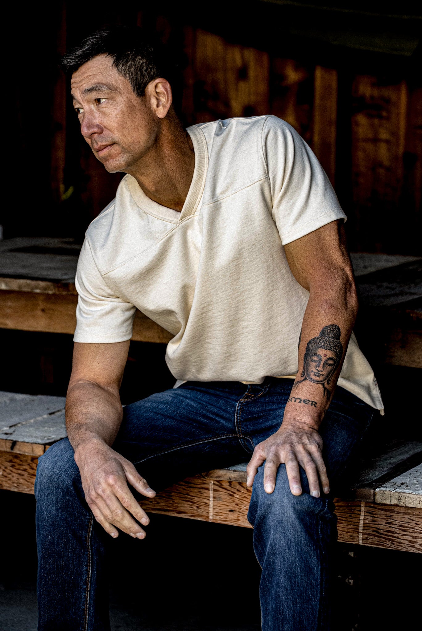 Man in a natural 12oz cotton V-neck jersey seated on weathered wood beside dark jeans.