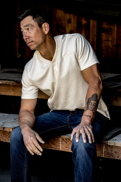 Man in a natural 12oz cotton V-neck jersey seated on weathered wood beside dark jeans.