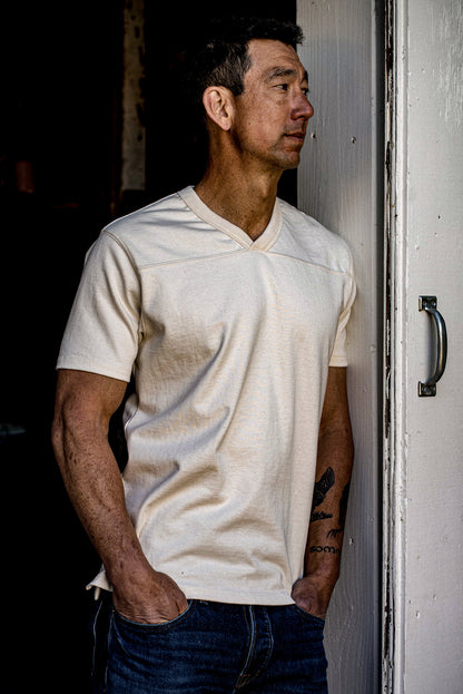 Man wearing natural Stinson V-neck cotton jersey, leaning against a weathered doorframe.
