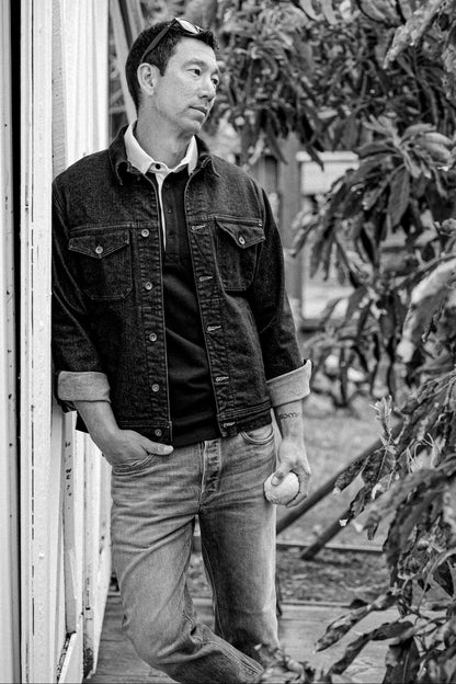 Man in a black broken twill denim jacket with rolled cuffs, leaning against a porch post in a black-and-white photo.