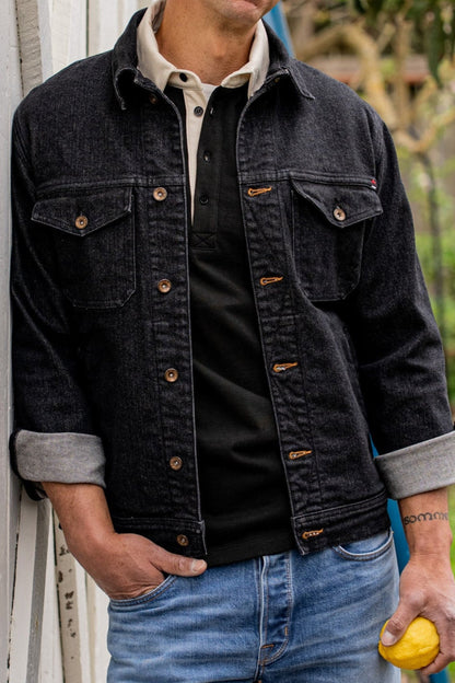 Black broken twill denim jacket with tobacco keyholes, brass buttons, and cuffed sleeves worn over a henley.