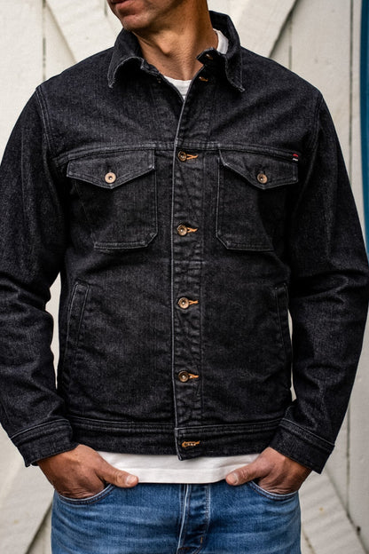 Front view of black broken twill denim jacket with chest pockets and brass buttons, worn over a white tee.