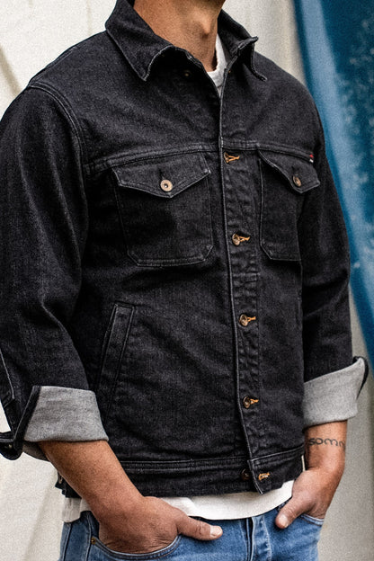 Model wearing black broken twill denim jacket with chest flap pockets, brass buttons, and cuffed sleeves.