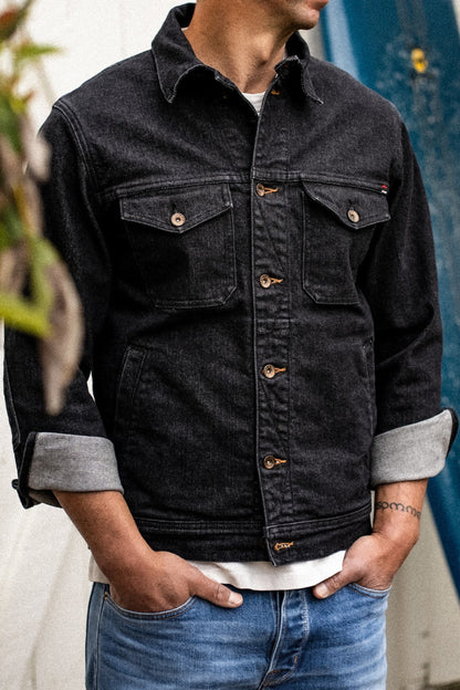 Model wearing black broken twill denim jacket with brass buttons, chest flap pockets, and cuffed sleeves over a white tee.