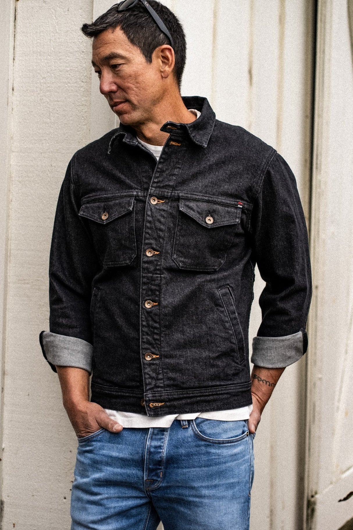 Man wearing a black broken twill denim jacket with brass buttons and cuffed sleeves over blue jeans.