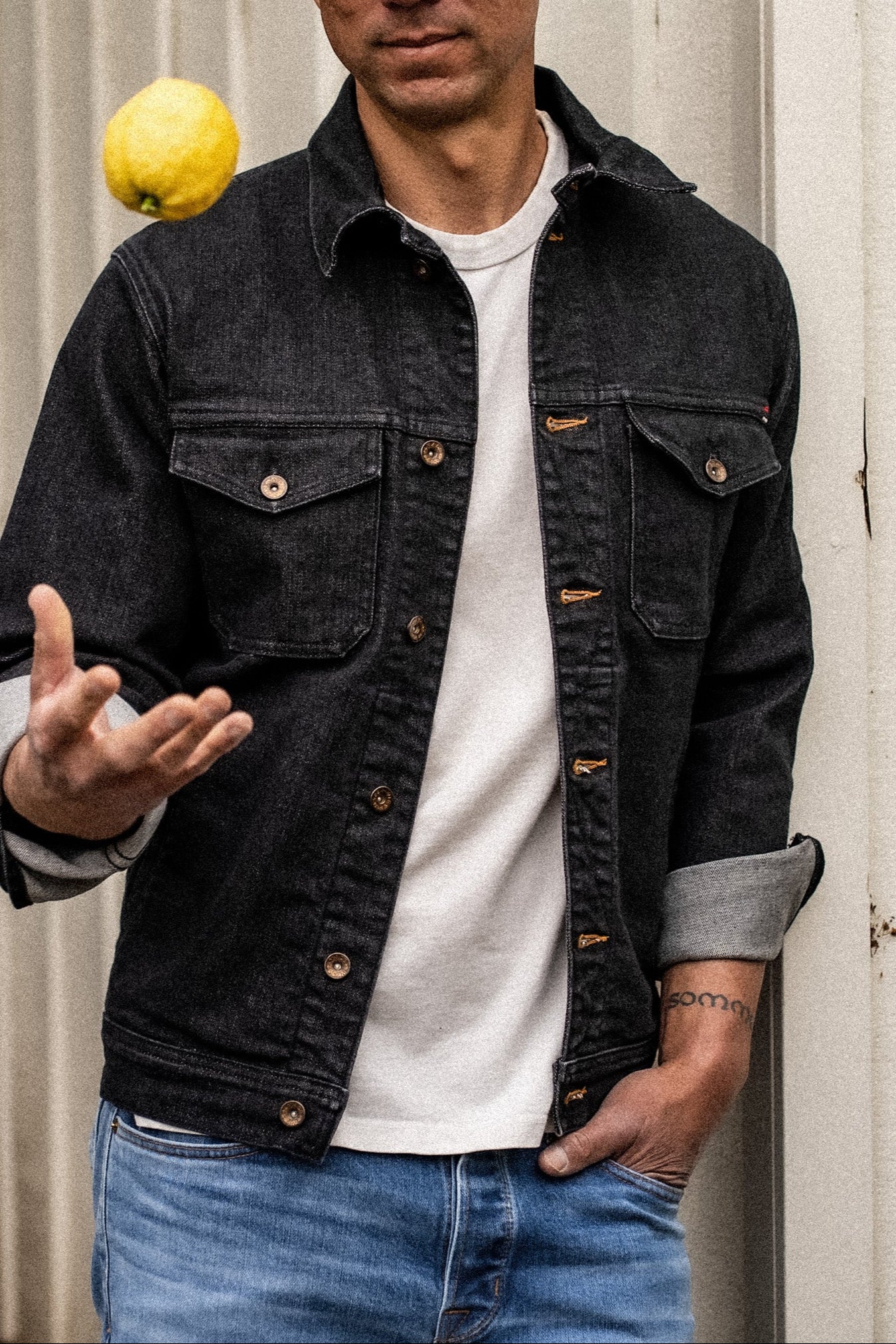 Man in black broken twill denim jacket with rolled cuffs and brass buttons, tossing a lemon.