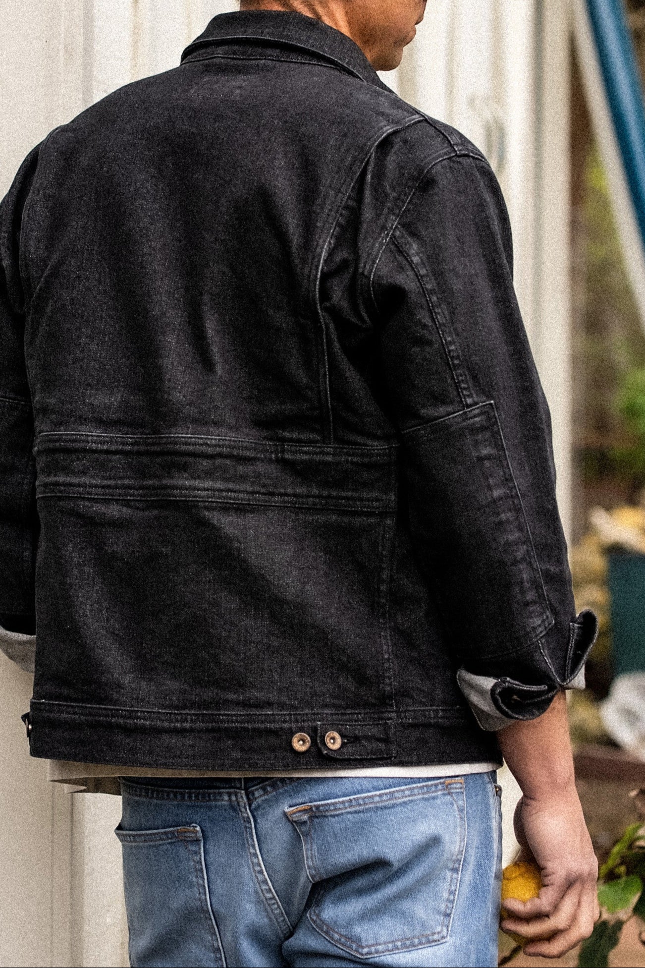 Back view of a black broken twill denim jacket with back gussets, brass buttons, and cuffed sleeves, worn with blue jeans.