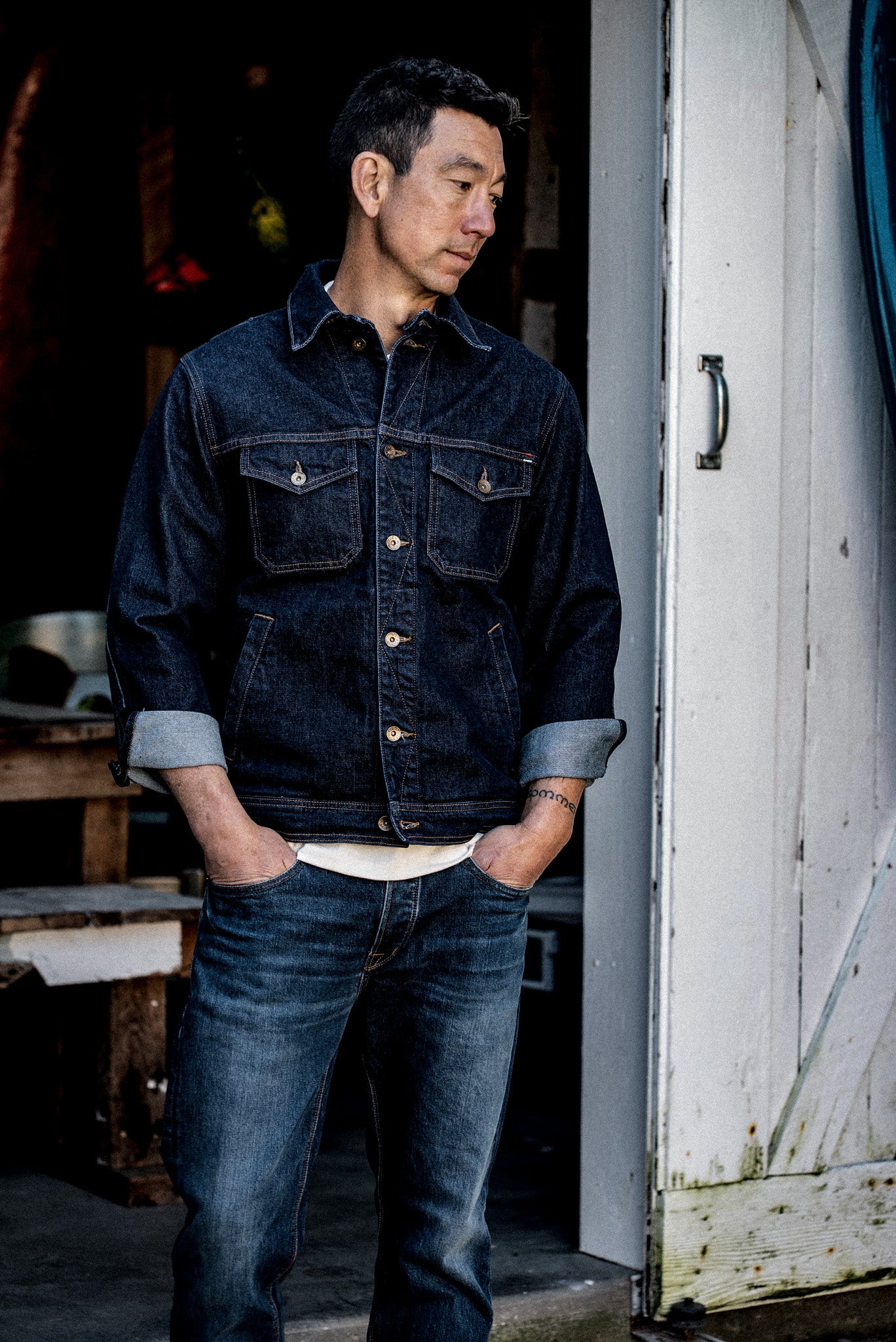Man wearing the Artcher broken twill denim jacket in ocean blue with rolled cuffs, standing in a doorway.