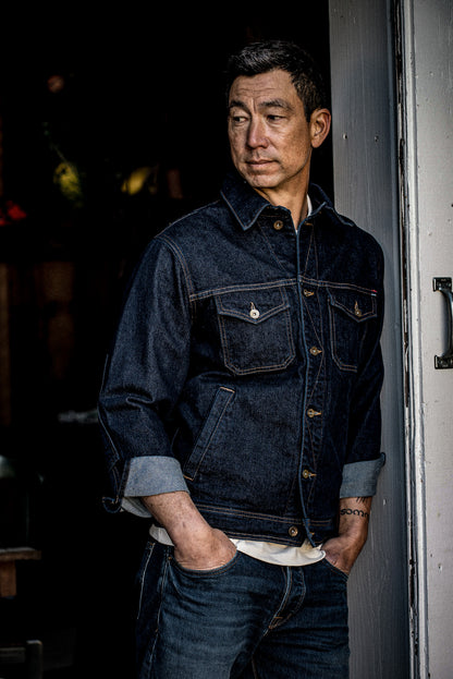 Man in dark indigo broken twill denim jacket with cuffs rolled to show selvedge, leaning in a doorway.