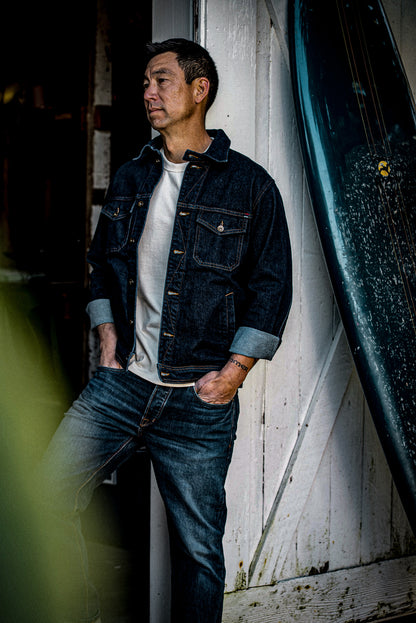 Man in dark ocean-blue broken twill denim jacket with cuffed sleeves, leaning against a weathered white door beside a canoe.