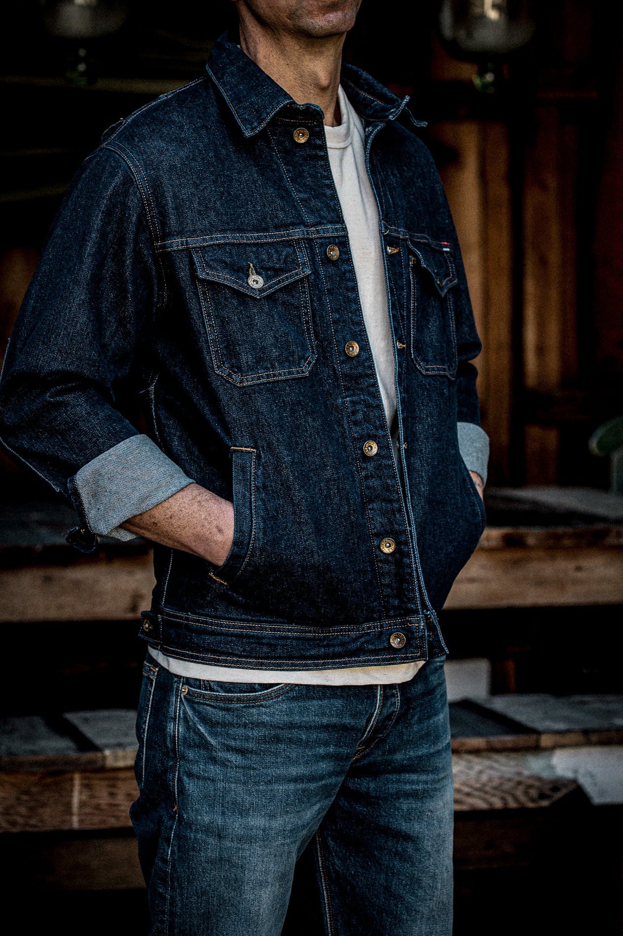 Man wearing dark indigo broken twill denim jacket with brass buttons and cuffed sleeves over a cream tee.