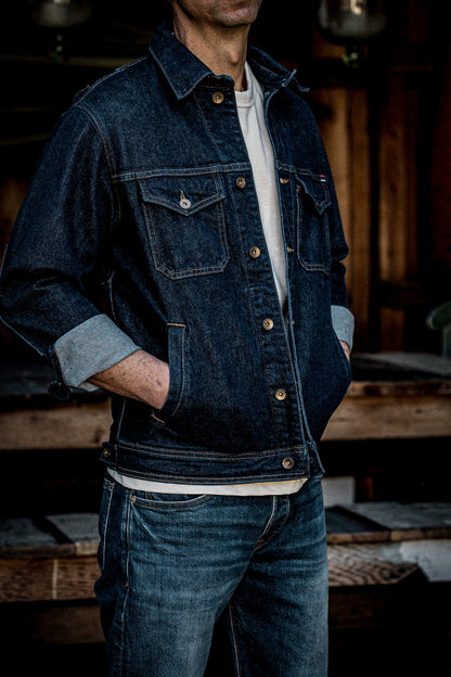 Man wearing dark indigo broken twill denim jacket with brass buttons and cuffed sleeves over a cream tee.