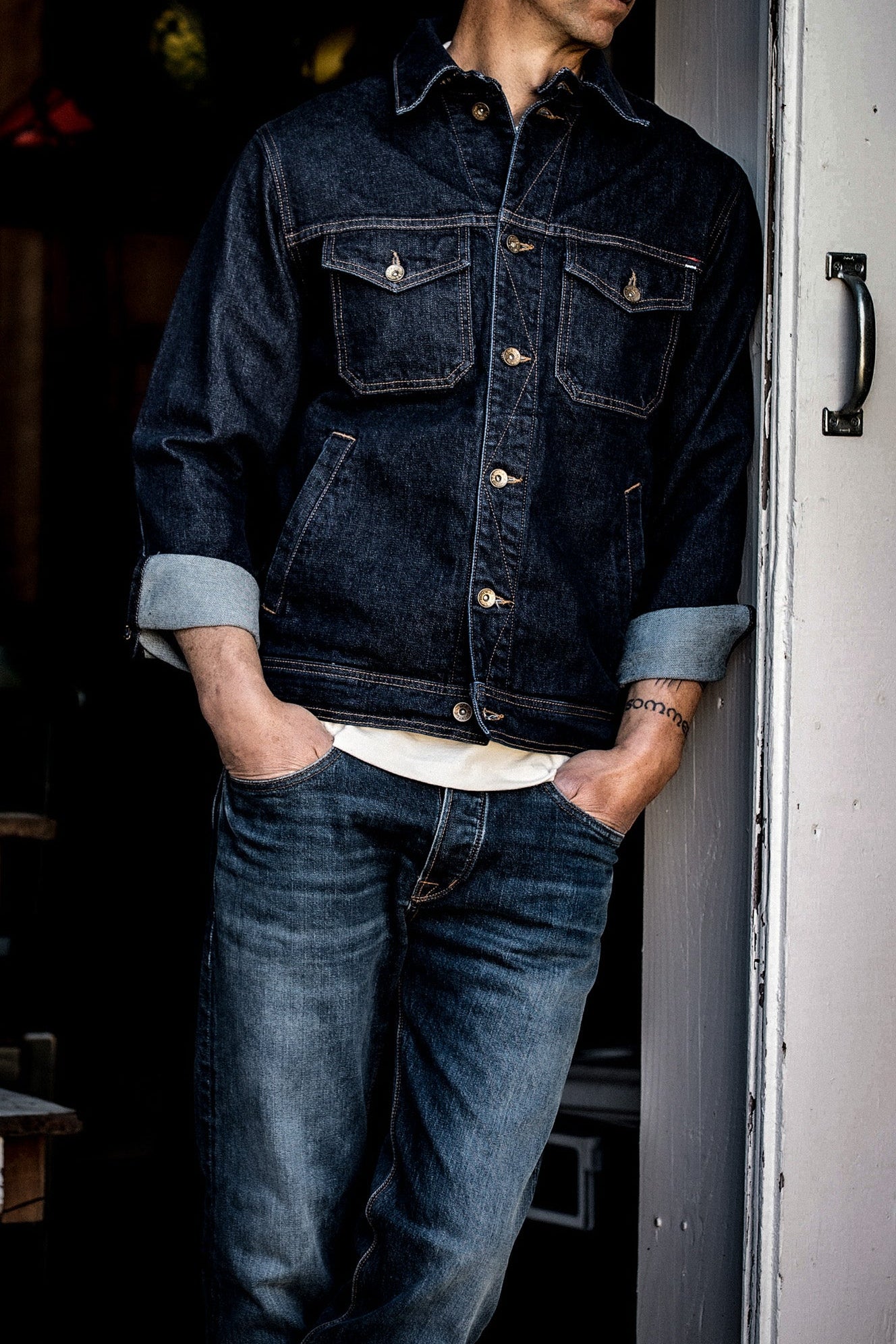 Person wearing Artcher ocean denim jacket with rolled cuffs, leaning in a doorway.